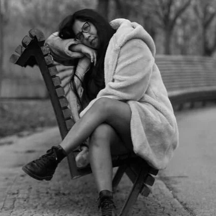A young woman with glasses sitting on a park bench, resting her head on her arm, in a cozy coat, with legs crossed, in an outdoor park setting.
