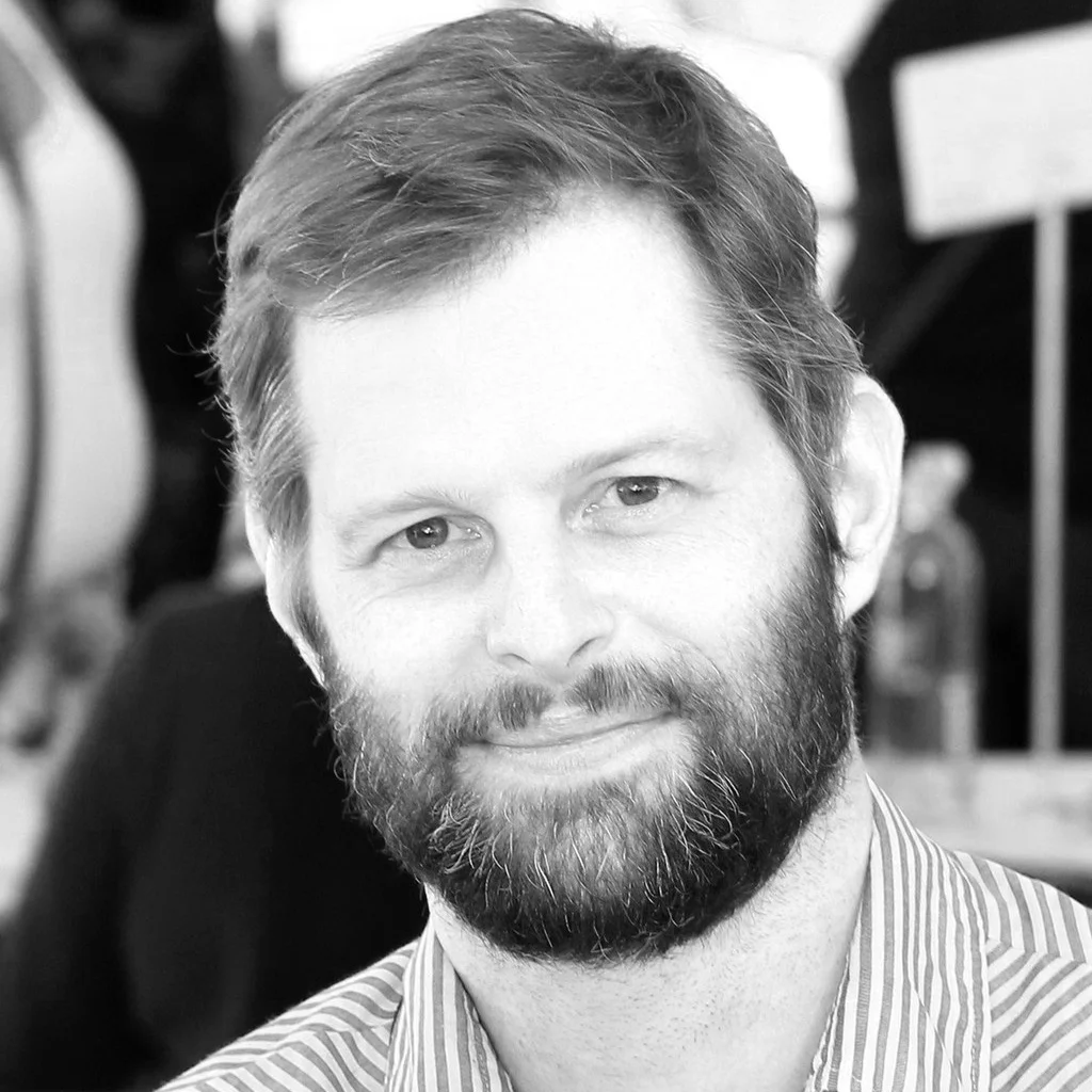 Black and white photo of a man with a beard and short hair, smiling, in an indoor setting.