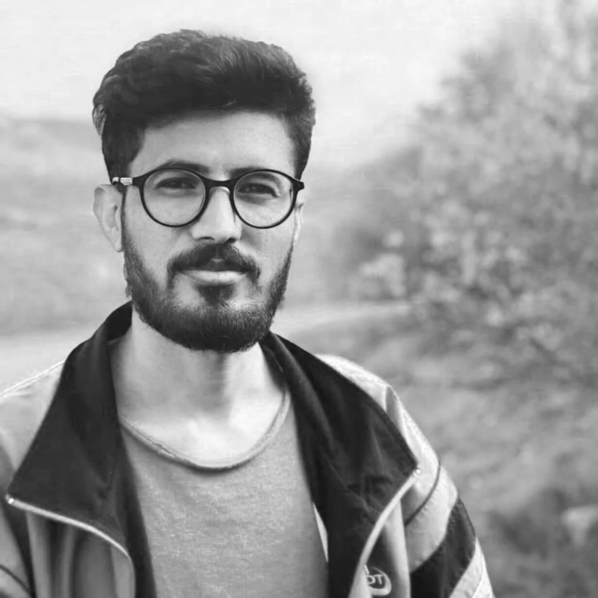 Black and white portrait of a man with glasses and a beard, outdoors with trees in the background.