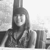 A woman with shoulder-length hair and bangs smiling, sitting indoors near a window.
