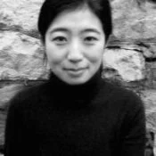 A woman with short hair wearing a black turtleneck, standing in front of a rock wall.