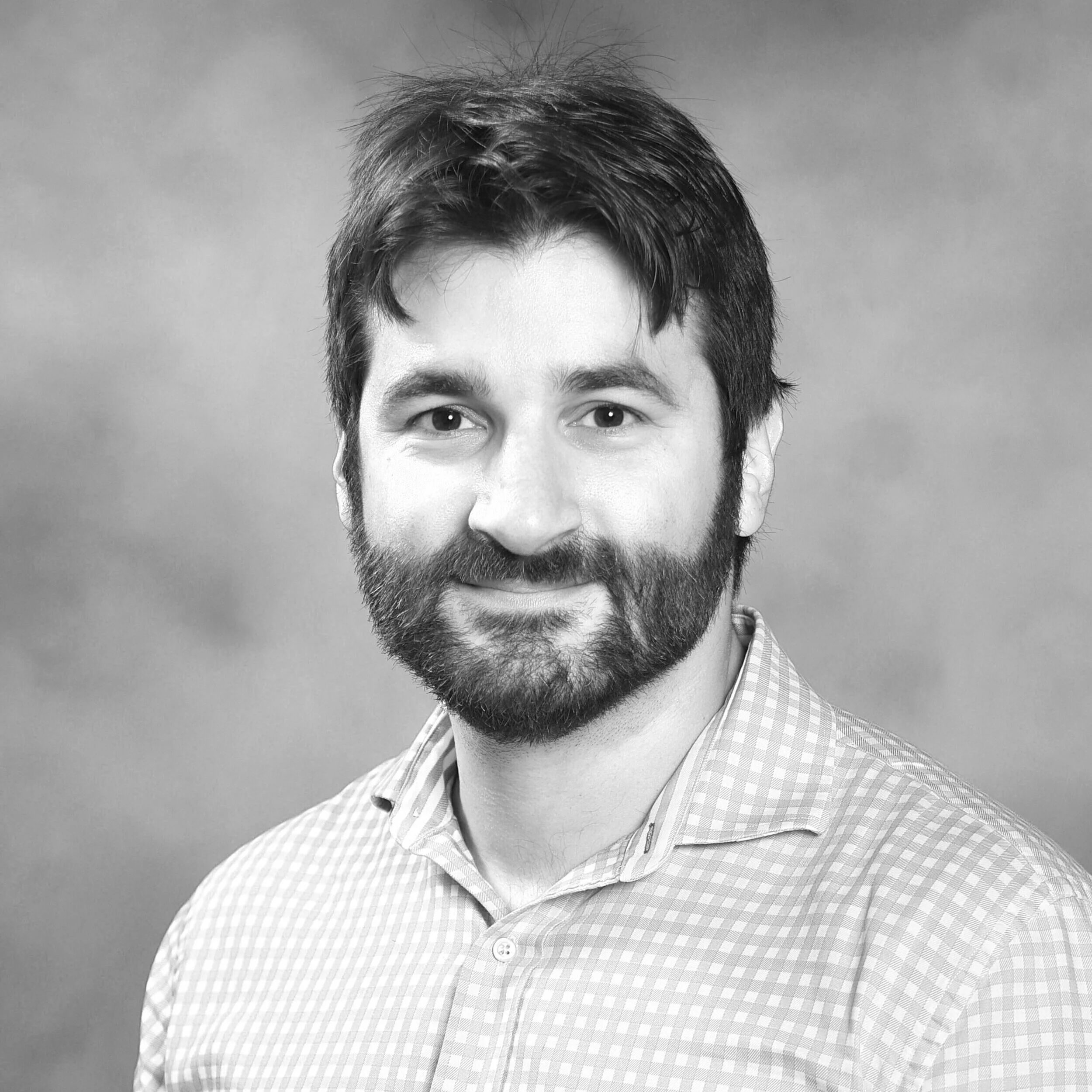 Black and white portrait of a man with a beard and short, slightly tousled hair, wearing a checkered collared shirt, smiling slightly, against a plain background.
