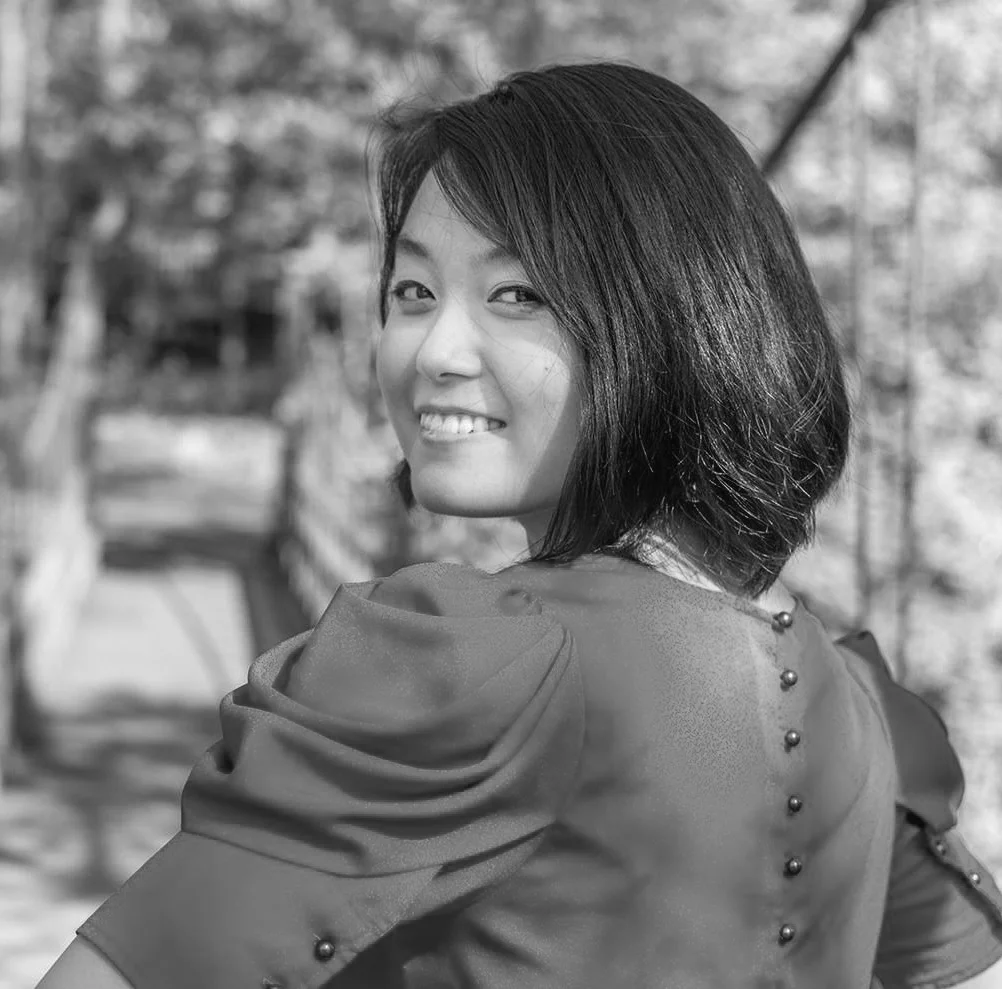 A woman with short dark hair smiling and looking back over her shoulder outdoors, wearing a puffed sleeve blouse.