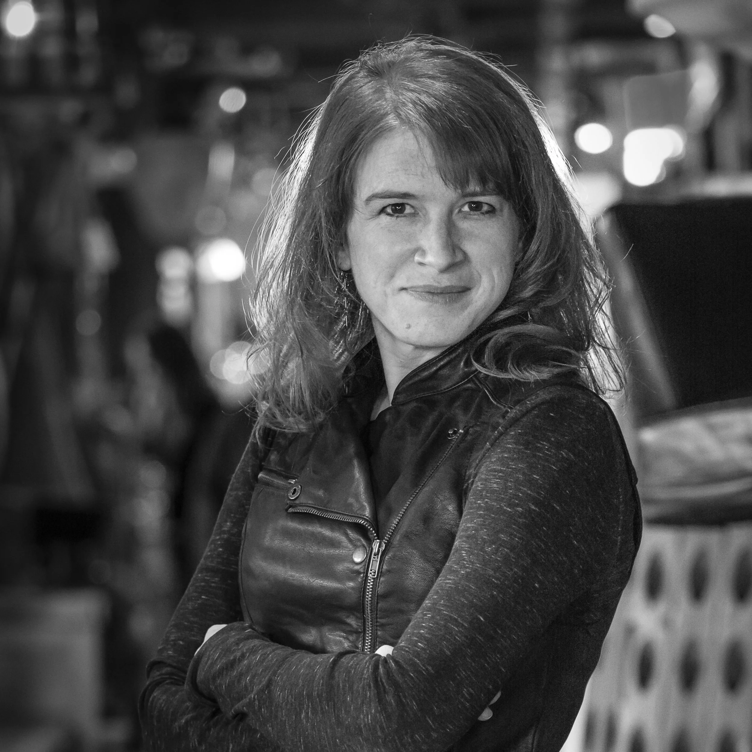 A woman with shoulder-length hair and a slight smile, wearing a dark leather jacket, standing with arms crossed. The background includes blurred lights and objects, suggesting an indoor setting.