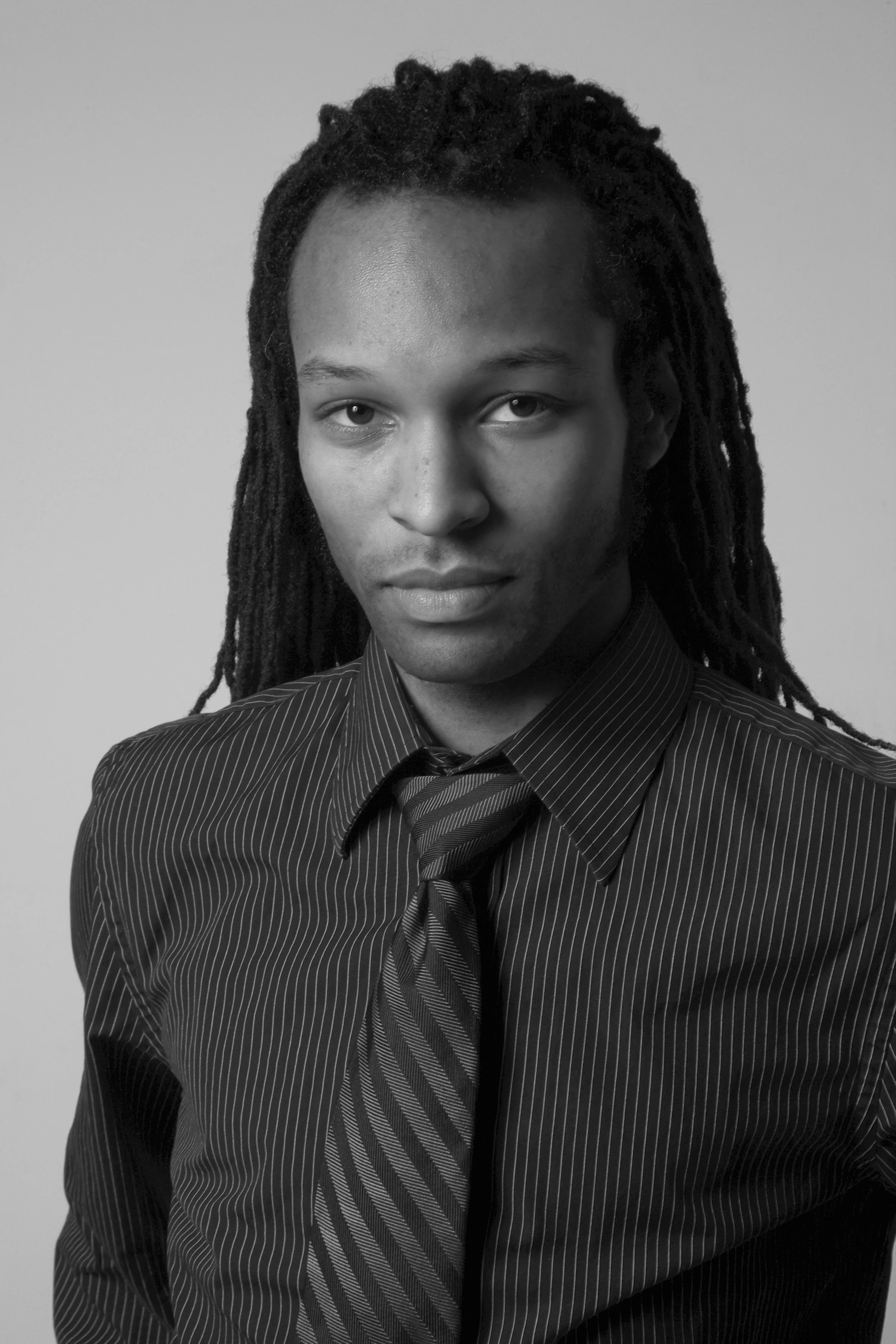 A black and white portrait of a man with long dreadlocks, wearing a pinstripe shirt and tie, looking directly at the camera.