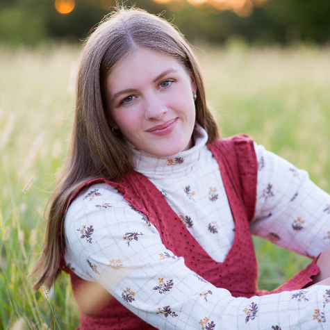 Young woman with long brown hair sitting in a grassy field, smiling at the camera, wearing a white blouse with a floral pattern and a maroon vest.