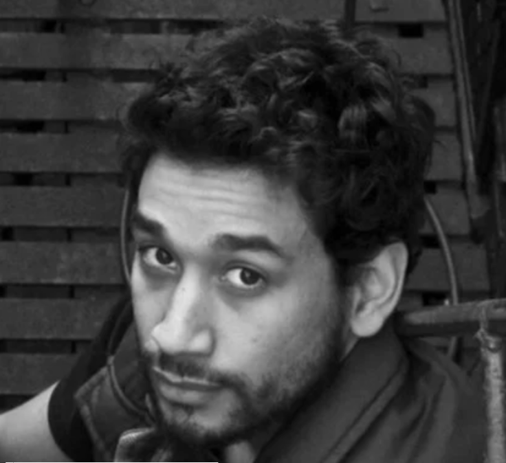 Young man with curly hair looking at the camera, sitting outdoors against a wooden fence.