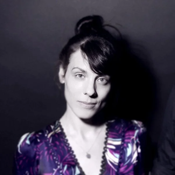 A woman with dark hair styled in a messy bun, wearing a colorful patterned top with purple and black hues, standing against a dark background.