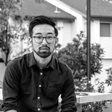 A man with glasses and a beard sitting outside near a house with trees and plants in the background.