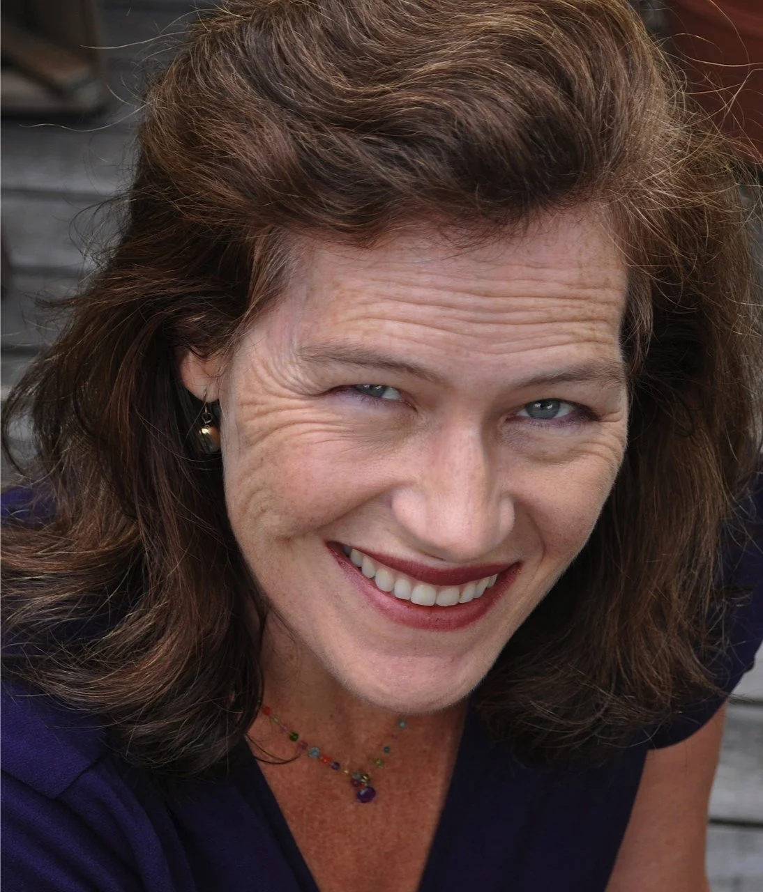 Close-up of a smiling woman with brown hair, blue eyes, wearing earrings and a colorful necklace.