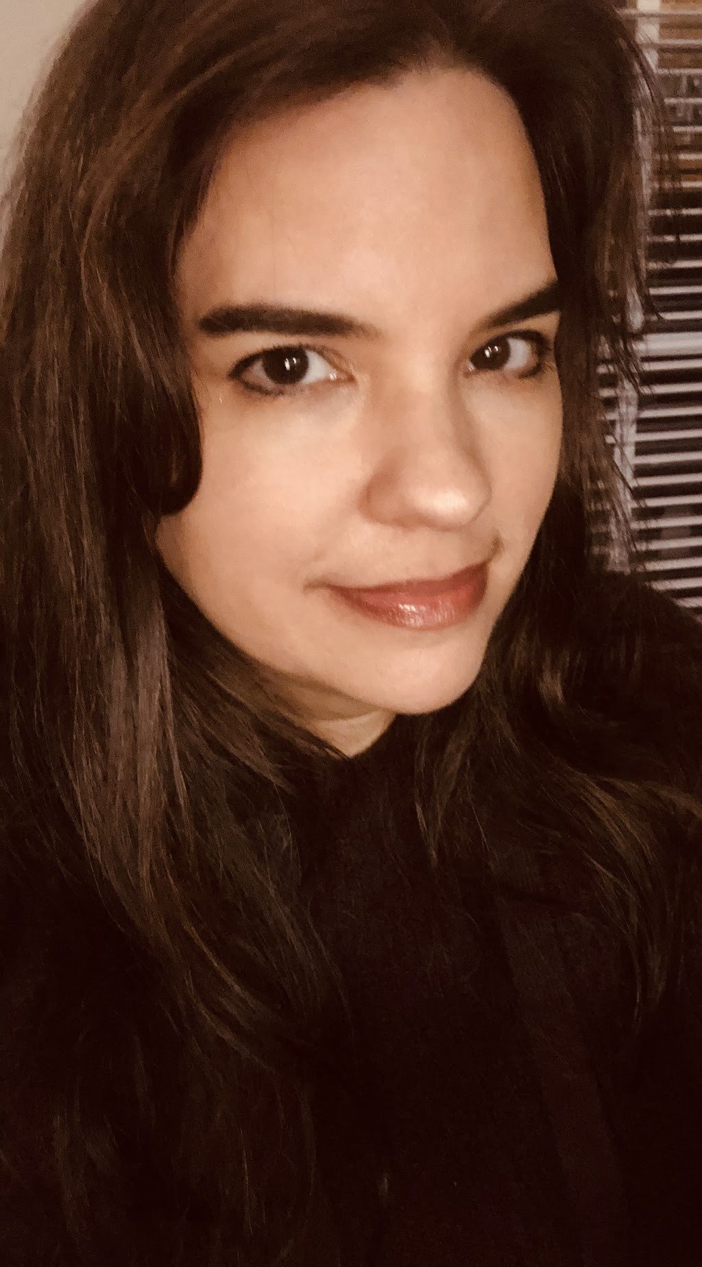 Close-up of a woman with long brown hair, wearing makeup with shiny lipstick, looking into the camera with a slight smile, background with wooden blinds.