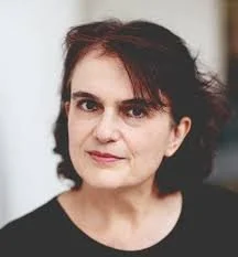 A woman with dark hair and fair skin, looking directly at the camera against a plain background.