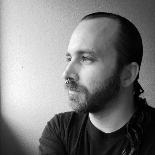 A black and white portrait of a man with long hair tied back, a beard, and a septum piercing, looking to the left.