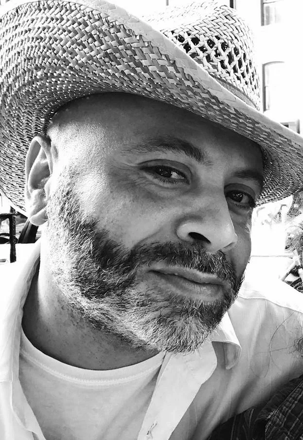 A man with a beard and mustache wearing a woven straw hat and a light-colored shirt, looking at the camera with a slight smile.
