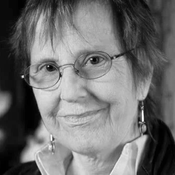 Close-up black-and-white portrait of an older woman wearing glasses and earrings, smiling gently.