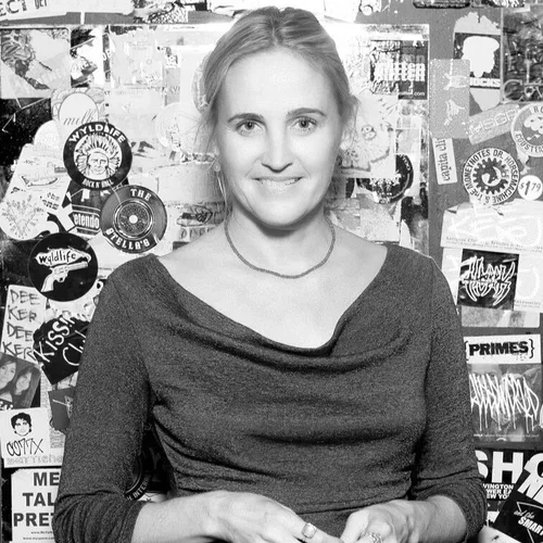 A woman smiling in front of a wall covered with stickers and posters.