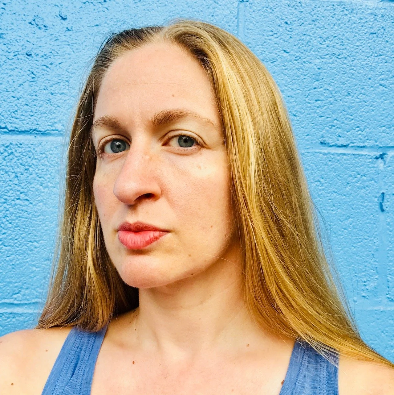 A woman with fair skin, blue eyes, and long blonde hair standing in front of a blue textured wall, wearing a blue tank top.