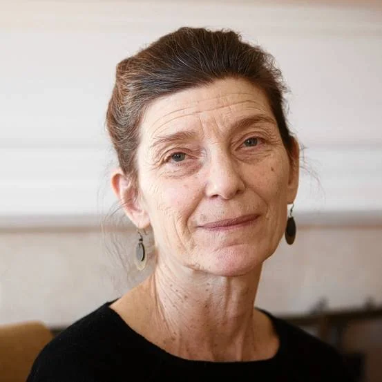Close-up of an older woman with brown hair tied back, wearing black earrings and a black top, sitting indoors with a white wall in the background.