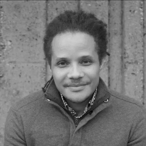 A black and white portrait of a man with curly hair, smiling, wearing a collared shirt and a zip-up jacket, standing in front of a textured wall.