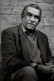Black and white portrait of an older man with gray hair, wearing a blazer and a collared shirt, seated outdoors in front of a brick wall.