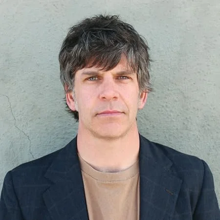 Headshot of a man with grayish-brown hair, wearing a black blazer over a tan t-shirt, standing against a gray wall.