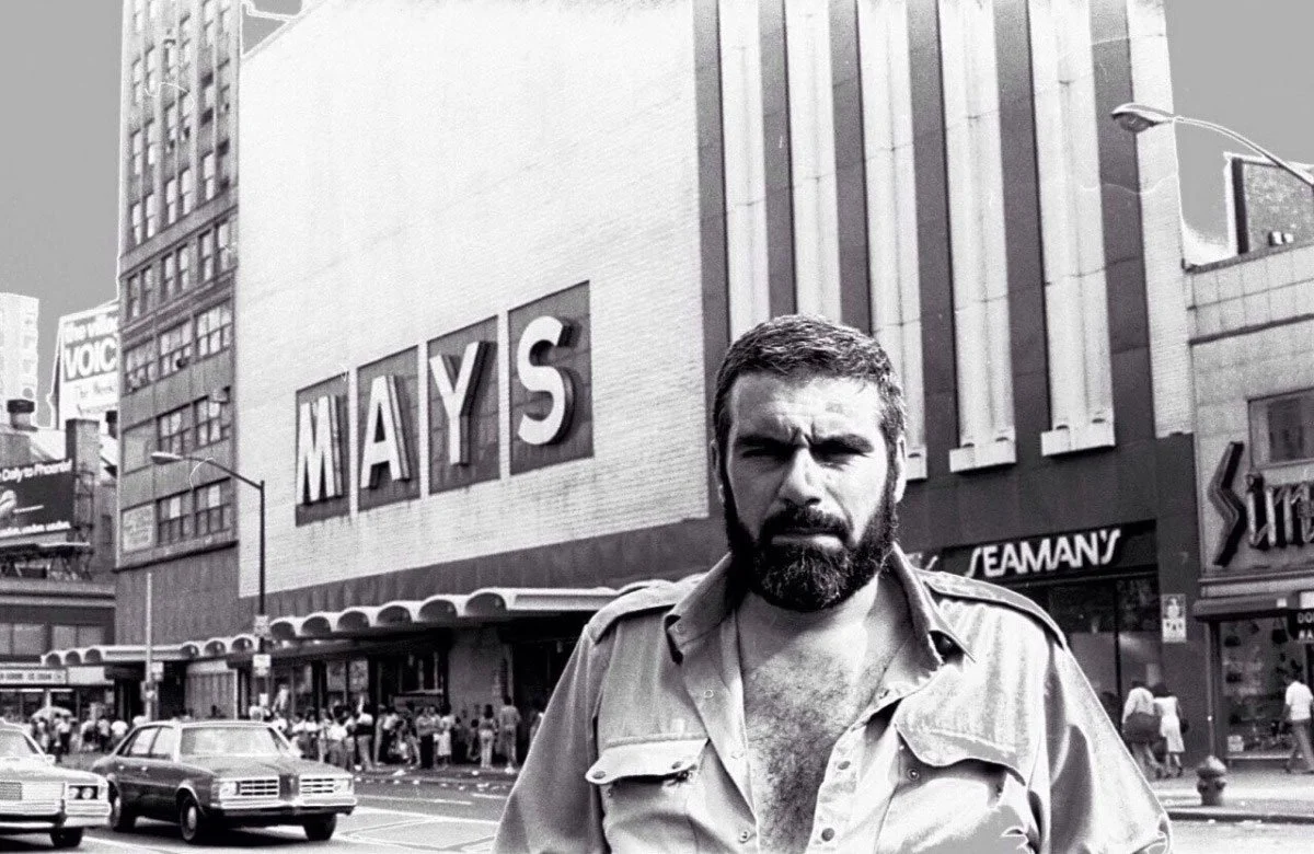 A man with a beard and open shirt standing on a city street in front of a large building with a sign that says MAYS, with cars and pedestrians in the background.