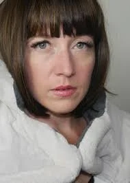 A woman with short brown hair and bangs looking directly at the camera, wearing a white top against a neutral background.