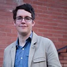A young man with dark curly hair and glasses, wearing a beige jacket and blue shirt, standing outdoors in front of a brick wall.