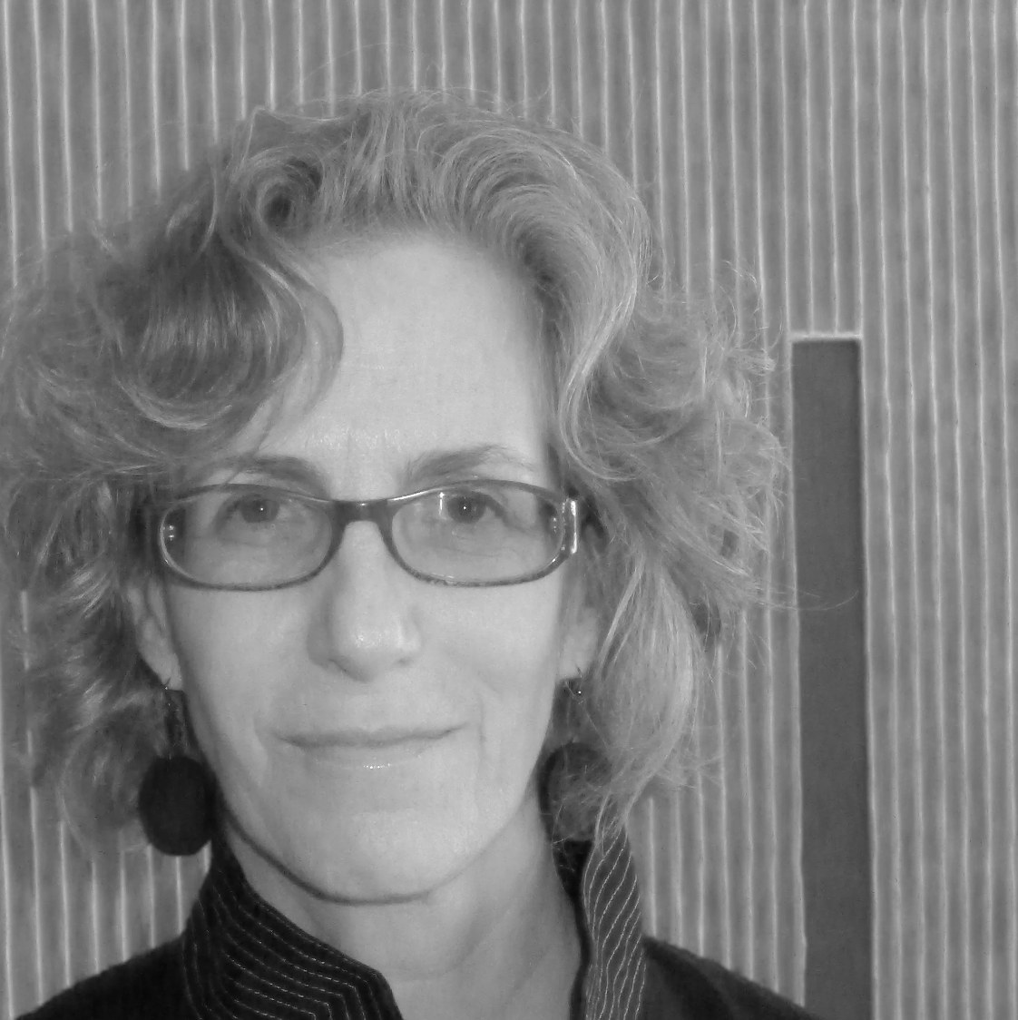 Black and white photo of a woman with curly hair, wearing glasses and earrings, smiling softly against a textured background.
