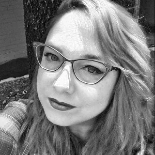 Black and white photo of a woman with glasses and long wavy hair, outdoors near a tree.