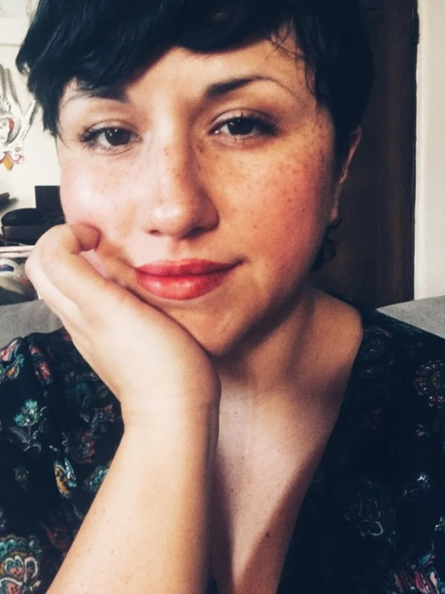 Close-up of a woman with short dark hair, freckles, and red lipstick resting her head on her hand, smiling softly.