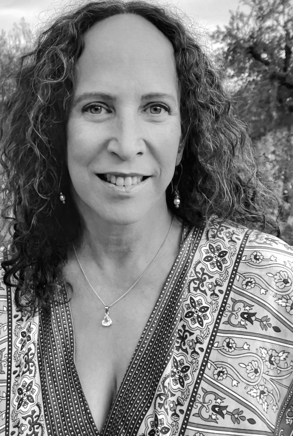 A woman with curly hair smiling outdoors, wearing earrings, a necklace with a pendant, and a patterned top.