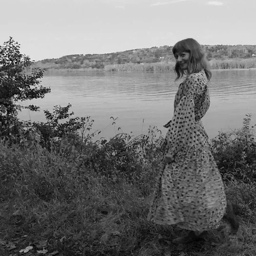 A woman in a polka dot dress standing near a lake with trees and hills in the background.