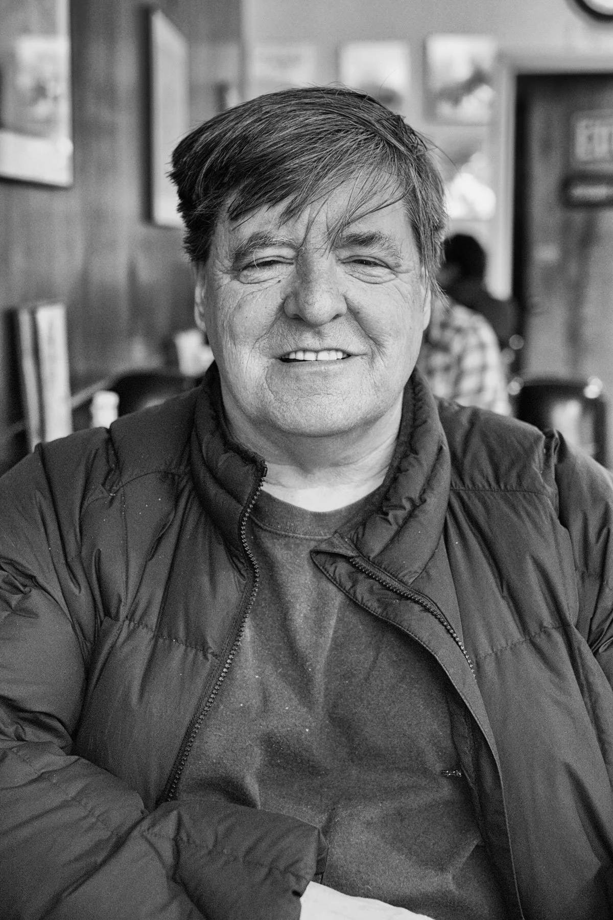 A black and white photo of a smiling man with medium-length hair, wearing a dark jacket over a sweater, sitting in a restaurant or cafe with framed pictures on the wall behind him.
