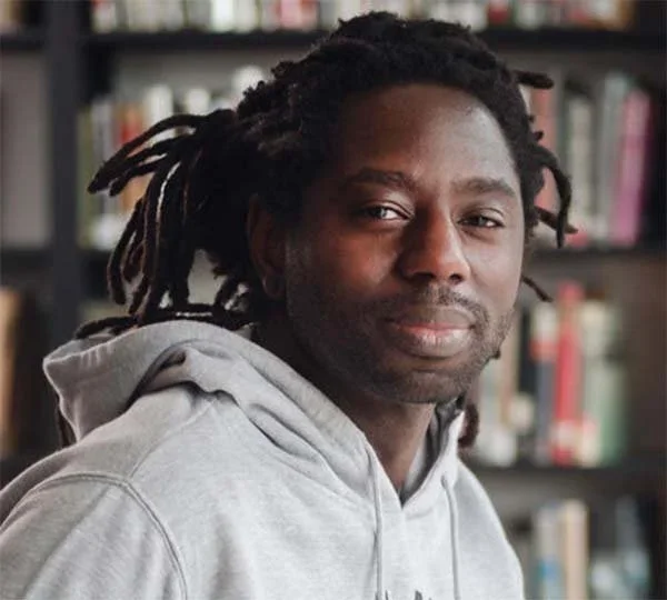 A man with dreadlocks, wearing a gray hoodie, standing in front of a blurred bookshelf.