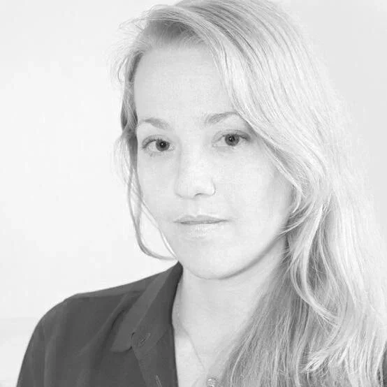 Black and white portrait of a woman with long, wavy hair, wearing a dark collared shirt, looking at the camera with a neutral expression.