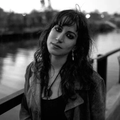 Black and white photo of a woman with dark hair and makeup, outdoors near a body of water.