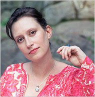A woman with dark hair wearing a pink patterned top, holding a pen near her face, outdoors with greenery in the background.
