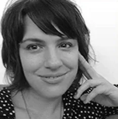 Black and white photo of a woman with dark hair smiling, wearing a polka dot top, resting her chin on her hand.