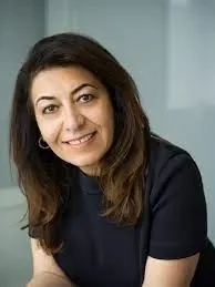 A woman with long dark hair smiling at the camera, wearing a dark top, in an office setting.