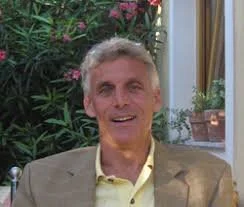 A smiling man with gray hair in a beige suit jacket and yellow shirt, sitting indoors with plants and curtains in the background.