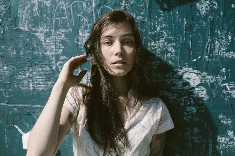 A young woman with long dark hair and light skin standing against a worn blue wall with white chalk markings, looking directly at the camera with a neutral expression.