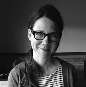A woman with glasses and dark hair smiling, wearing a striped shirt and a dark cardigan.