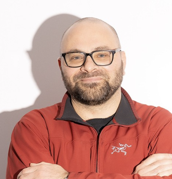 A man with glasses and a beard, wearing a red jacket with a black collar, standing with arms crossed against a white background.