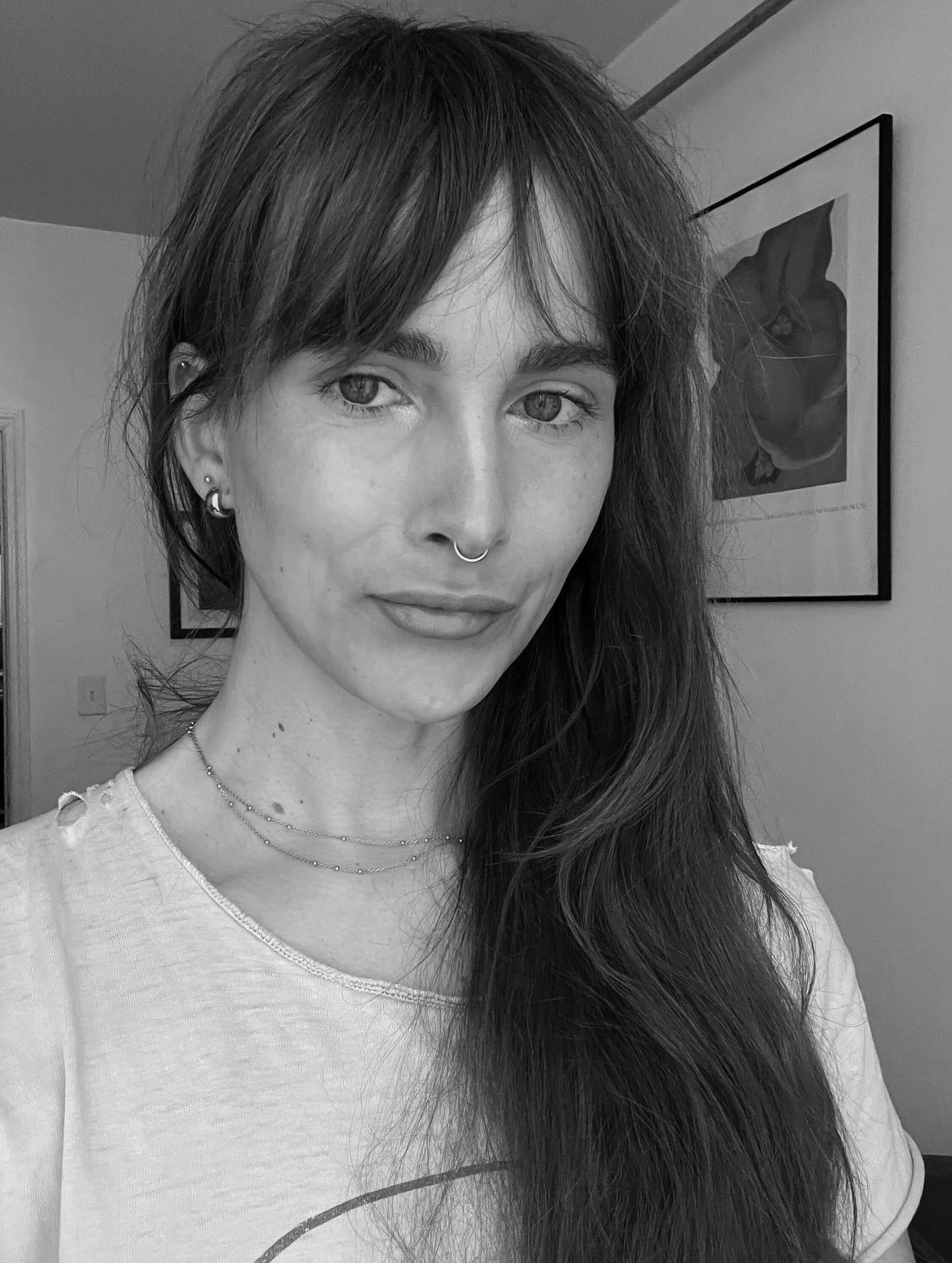 A woman with long hair and a septum piercing, looking at the camera with a neutral expression, standing in a room with towels and clothes in the background.