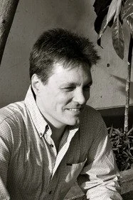 Black and white photo of a man smiling, sitting near indoor plants.