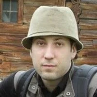 A person wearing a beige bucket hat and a dark jacket with a backpack in front of a wooden background.