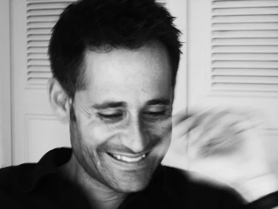 A smiling man with short dark hair, wearing a dark shirt, looking down and slightly to the side, with a blurred background.