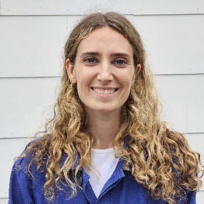 A headshot of a light-skinned woman with light curly hair in front of a white wall wearing a blue shirt.