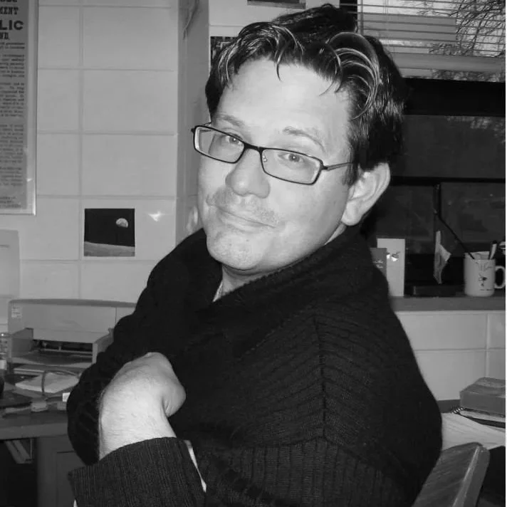 BW man sitting in chair, turned around to look at camera, straight dark cropped hair, glasses, dark button-up, smiling 